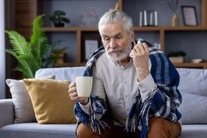 Sad senior gray-haired man sitting on sofa at home and suffering from illness. Covered with a blanket, holding a cup of hot drink and a napkin. photo