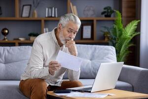 Problems with accounts and documents. Pensive and upset senior man sits at home on the sofa and looks sadly at the papers in his hands. photo