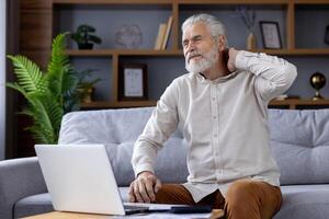 Tired senior gray-haired man sits at home on the sofa in front of the table with a laptop and holds his neck with his head, doing asana, resting. photo