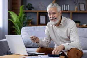 Portrait of smiling senior gray-haired man sitting on sofa at home and checking bills and documents, using laptop and calculator, looking at camera. photo