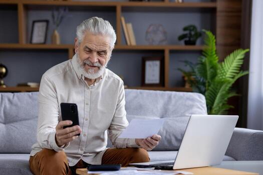 Smiling senior gray-haired man sitting at home on the sofa in front of the table with a laptop, holding a notebook and using a mobile phone. photo