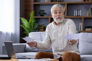 Portrait of a worried senior gray-haired man sitting on a sofa at home and working on a laptop, checking bills and received receipts, looking upset at the camera and throwing up his hands. photo