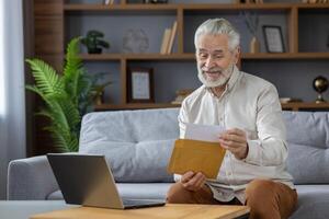 Senior gray-haired smiling man sits on a sofa and opens an envelope with a letter. Reads the received message, bill, invitation. photo