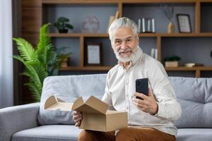 Portrait of a smiling gray-faced senior man who is happy to shop online, holds a delivered package, a mobile phone and smiles at the camera. photo