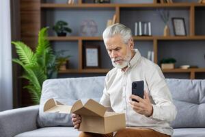 Shocked senior gray-haired man sits on the sofa at home, holds the phone in his hands and looks sadly at the open package he received in the mail. photo
