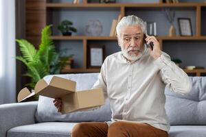 An older gray-haired man sits at home on the sofa and angrily talks on the phone, stubbornly holding in his hands an open package delivered by mail. photo