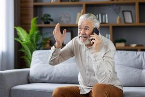Upset and angry senior man talking on the phone at home, sitting on the sofa and expressively explaining and gesturing with his hands. photo