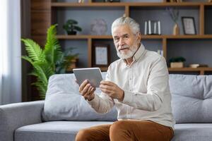 Senior gray-haired man sitting on the sofa at home, using a tablet, talking on a call and looking at the camera. photo