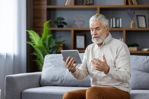 Senior gray-haired man communicates via call from a tablet with a doctor, conducts an online consultation, explains the problem, sitting on the sofa at home. photo