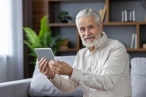 Close-up portrait of an older gray-haired man sitting on the couch at home, holding a tablet and smiling at the camera. photo