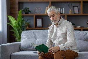 Gray-haired senior man sitting on the sofa at home and reading a book with concentration, resting, spending free time in retirement. photo