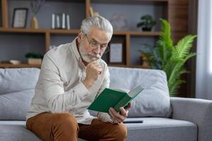 An elderly gray-haired man is sitting on the couch at home in an apron and is reading an interesting book with concentration. photo
