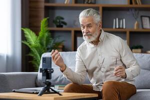 Senior gray-haired man sits on the sofa at home and talks on a call from the phone, records a podcast, conducts an online lecture. photo