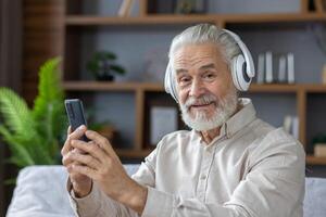 Close-up portrait of a senior gray-haired man sitting on the sofa at home wearing headphones and talking on a call from the phone, smiling and looking at the camera. photo