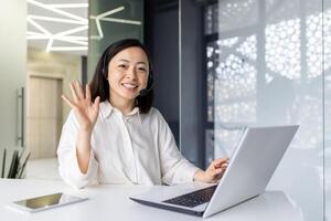 Portrait of an Asian young woman working in the office using a laptop, sitting at a desk in a headset, consulting and talking online, smiling and waving at the camera. photo