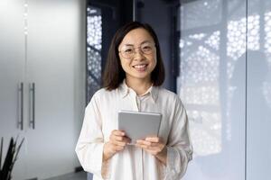 Portrait of successful asian female programmer inside office, young woman smiling and looking at camera, using tablet computer to test application software, workplace inside office. photo