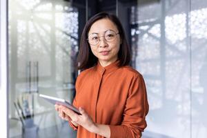 Portrait of a young Asian woman programmer, developer, tester in glasses standing in the office, holding a tablet, working online and looking, posing at the camera. photo