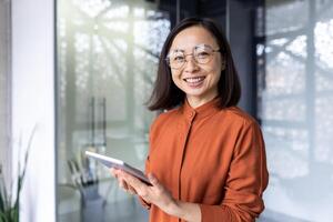Portrait of a young successful Asian female programmer inside office, worker in glasses smiling and looking at the camera, a businesswoman holds a tablet computer in hands and uses it to test online . photo
