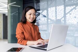 Successful asian business woman working inside office building with laptop, female worker smiling and looking at computer screen, satisfied with work achievement results. photo