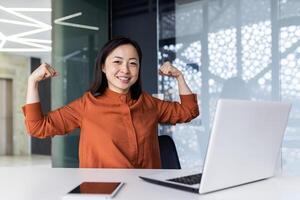 un joven asiático negocio mujer trabajando en el oficina utilizando un computadora portátil, con confianza y sonriendo mirando a el cámara y demostración un poder gesto con su manos. exitosamente terminado trabajar, proyecto. foto