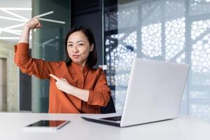 Portrait of a young successful Asian female programmer, developer and IT expert. Sitting at a table and working on a laptop, pointing at the camera with a finger on his hand, a gesture of strength. photo