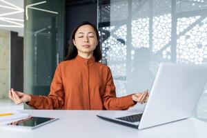 Young beautiful Asian web developer working inside office building, woman resting meditating at workplace with eyes closed, female worker using laptop to write code photo