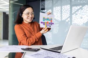 A cheerful Japanese woman in a headset sits at a table in a modern office, talks on a call on a laptop, shows graphs to a webcam, conducts a seminar, shows a report. photo