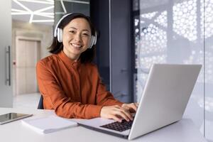 Young successful female programmer at workplace inside office working with laptop, coding software, smiling and looking at camera, female worker with headphones online music and podcasts. photo