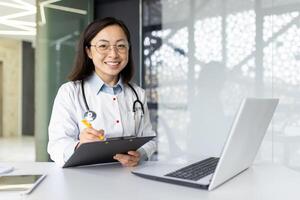 Portrait of a smiling Asian female doctor working in a hospital using a laptop, writing documents and case histories, consulting remotely and talking via call. photo