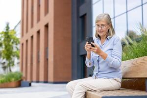 An older woman with glasses using a smartphone while sitting on a bench in an urban setting, near a modern building. photo