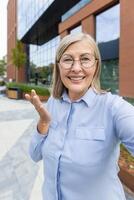 An attractive senior woman in glasses smiles and gestures while taking a selfie outside near a modern building. photo