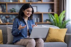 A smiling woman is seen using a laptop while sitting on a sofa, engaged in a call with friends or family. The scene suggests remote work or communication. photo