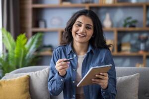 A smiling young woman holds a pen and notepad, possibly preparing to take notes or brainstorming ideas in a comfortable setting. photo