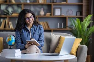 Distance learning and studying. Young smiling Indian woman sitting on sofa at home, talking on call and taking notes in notebook. photo