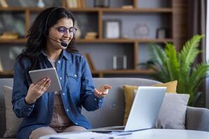 A Muslim woman in a headset sits at home on a sofa in front of a laptop, holds a notepad in her hands and talks via call to the camera. photo