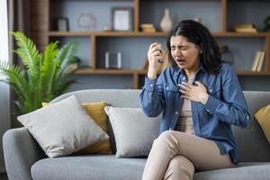 Young Indian woman suffering from asthma and allergies sitting on sofa at home, holding her hand on her chest and using inhaler. photo
