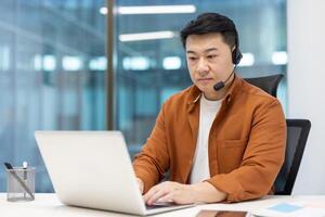 An Asian man with a headset uses a laptop in a modern office setting, showcasing remote work and customer service. photo