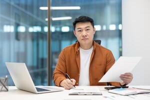 A focused Asian man reviews documents in a modern office setting, captured with natural light and a serious expression. photo