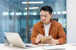 An Asian man sits at his desk, concentrating while taking notes and using his laptop in a modern office setting. photo