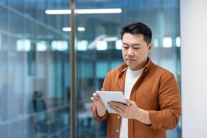 An Asian man in an office setting using a tablet. The background has glass windows. photo