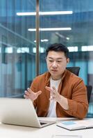 An Asian man in a business casual setting gestures while looking at a laptop, possibly during a call. photo