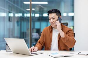 An asian man in a modern office talks on the phone while working on a laptop. Focus on communication and business. photo