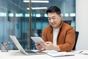 An Asian man smiles while working on a tablet and laptop at his desk in a modern office setting. photo