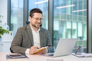 A smiling businessman works on a laptop in a modern office, writing in a notepad with a pen. Modern business environment. photo