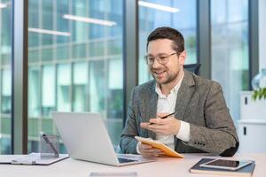 Smiling young businessman in a suit sitting at his workplace in the office and talking remotely via laptop, making notes in a notebook. photo