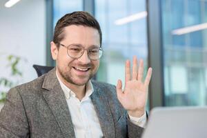 A smiling businessman waves during a call, working in a modern office environment. photo