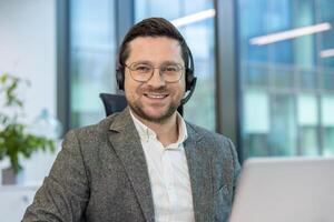 Portrait of a young man in a headset and suit sitting in an office at a desk with a laptop and looking at the camera with a smile. photo