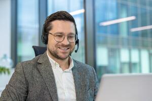 A close-up photo of a young man in a suit and headset sitting in an office at a desk and working on a laptop with a smile.
