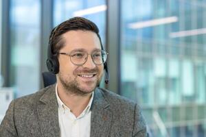 A close-up photo of a young man wearing a headset and glasses who is working in an office, smiling and looking to the side.