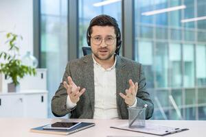 Portrait of a young man in a headset and computer sitting in an office at a table and holding an online conference, explaining by gesturing with his hands. photo